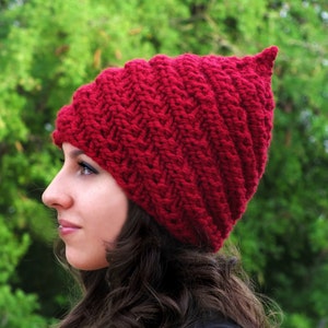 Op de afbeelding: Een rode gebreide muts met een puntige top. De muts is gemaakt met een chevron-steekpatroon.