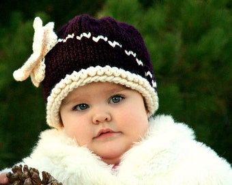 Puede incluir: Un bebé con un abrigo de piel blanco y un gorro de punto morado y blanco con un detalle de flor. El gorro tiene un borde blanco y un cuerpo de punto morado con costuras en zigzag blancas.