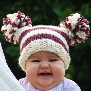 Peut inclure: Un bébé portant un bonnet tricoté rayé blanc et violet avec des pompons blancs sur le dessus. Le bébé sourit et a un visage rond.