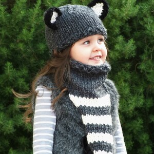 May include: A young child wearing a gray and white striped knit scarf and a gray knit hat with black ears. The hat and scarf are handmade and have a cozy, wintery feel.