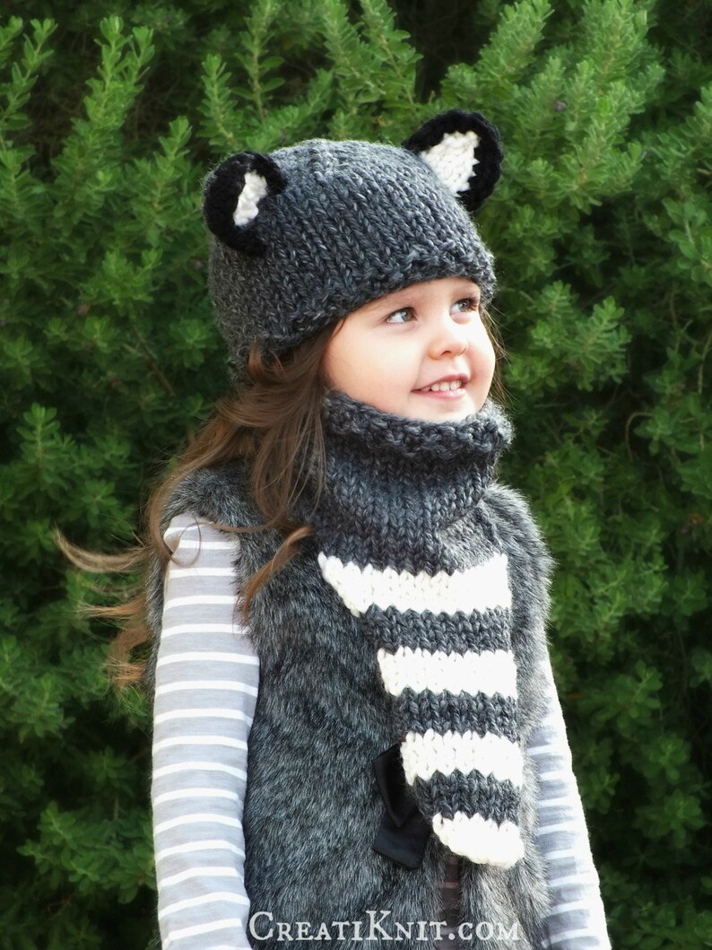 May include: A young child wearing a gray and white striped knit scarf and a gray knit hat with black ears. The hat and scarf are handmade and have a cozy, wintery feel.