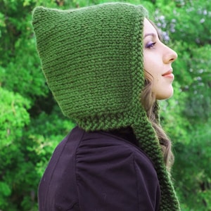 May include: A woman wearing a green knitted hooded scarf. The scarf is long and has a pointed hood. The woman is wearing a black jacket and has long brown hair.