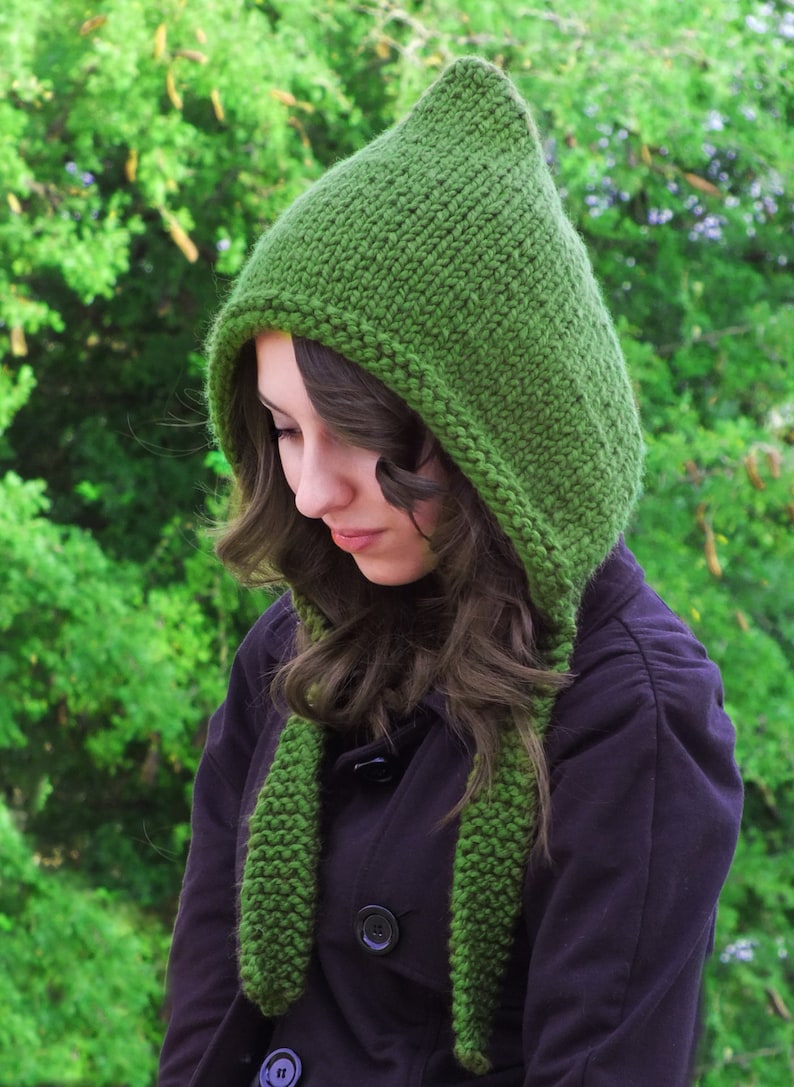 May include: A green knitted hooded scarf with a long, flowing design. The scarf is worn by a person with long brown hair.