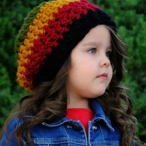 Puede incluir: Una niña pequeña con un gorro de punto de ganchillo de colores con rayas verdes, amarillas, rojas y negras. Lleva una camiseta roja y una chaqueta vaquera azul.