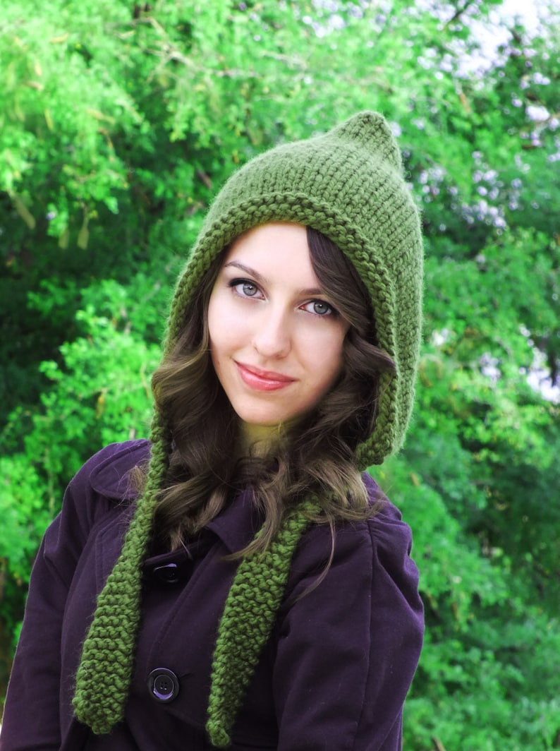 May include: A woman wearing a green knitted hooded hat and a dark purple jacket. The hat has a long scarf attached to it.