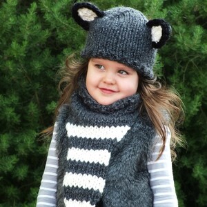 May include: A gray knitted hat with black ears and a white and gray striped scarf. The hat and scarf are worn by a child.