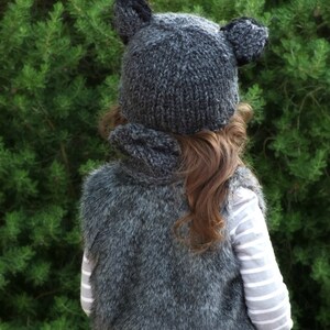 May include: A gray knitted hat with bear ears and a matching gray faux fur vest. The hat and vest are worn by a child.