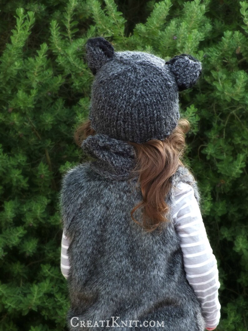 May include: A gray knitted hat with bear ears and a matching gray faux fur vest. The hat and vest are worn by a child.