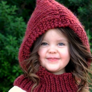 Puede incluir: Un niño pequeño con un gorro de punto rojo con capucha y una bufanda a juego. El gorro tiene una punta puntiaguda y la bufanda es de estilo cuello alto.