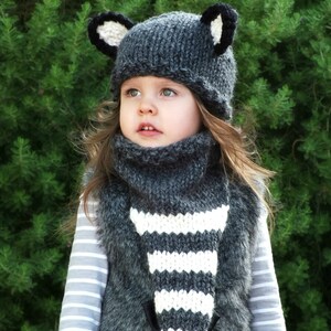 May include: A gray knitted hat with black ears and a matching gray and white striped scarf. The hat and scarf are worn by a young child with long brown hair.