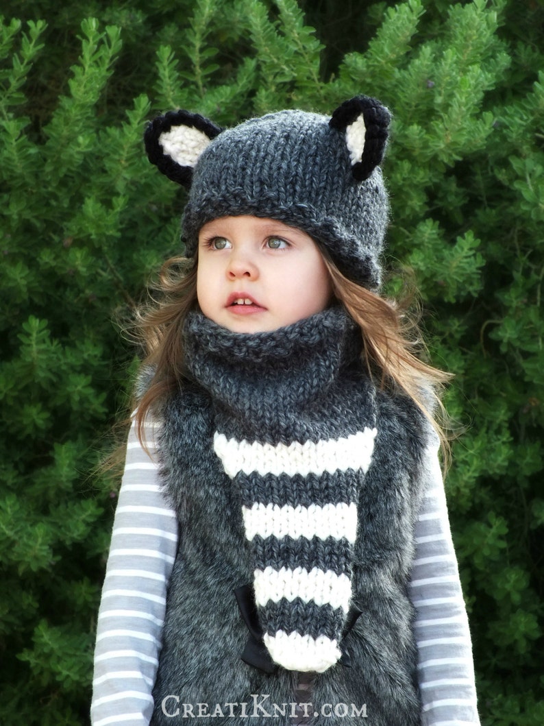 May include: A gray knitted hat with black ears and a matching gray and white striped scarf. The hat and scarf are worn by a young child with long brown hair.