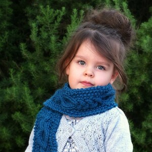 May include: A young girl wearing a teal blue knitted scarf with a cable knit pattern. The scarf is wrapped around her neck and tied in a loose knot. She is wearing a light gray sweater with a white lace overlay. The girl has brown hair pulled back in a bun. She is standing in front of a green leafy background. The text "CREATIKNIT.COM" is visible at the bottom of the image.