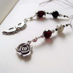 May include: A silver chain necklace with a silver rose charm, a silver mask charm, and red and black beads.