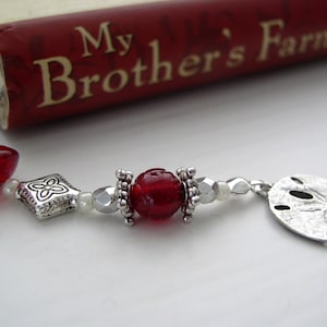 May include: A red and silver bookmark with a sand dollar charm. The bookmark has red glass beads, silver beads, and a silver charm with a floral design. The bookmark is attached to a book titled "My Brother's Farm".