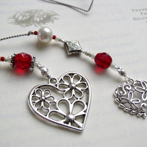May include: Two decorative bookmarks with heart-shaped charms. One charm is a large openwork heart with flowers, the other is a smaller openwork heart. Both have red and white beads.