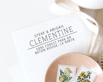 May include: A white card with the name "CLEMENTINE" and address details in black font. Three floral stamps rest on a pink plate. A white ribbon and a white cup are in the background.