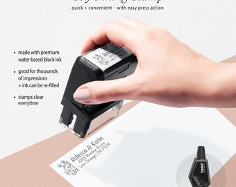 May include: A black self-inking stamp with a floral design and the text "Rebecca & Kevin 4285 Meadow Brooks Lake Oswego, OR 97034". The stamp is being held by a hand. A small bottle of black ink is shown in the bottom right corner with the text "Re-Fill Ink available as option".