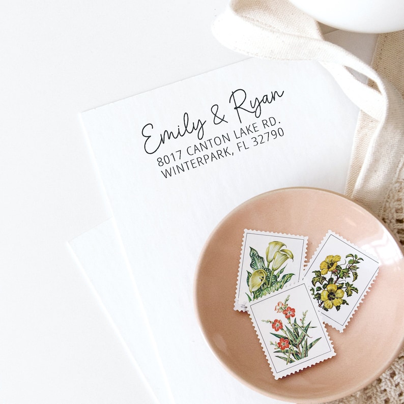 May include: A white envelope with a black return address stamp that reads "Emily & Ryan 8017 Canton Lake Rd. Winterpark, FL 32790".  A pink saucer with three vintage floral stamps is on top of the envelope.