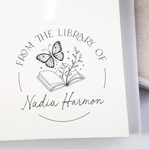 May include: A white card with a black ink illustration of a butterfly, open book, and foliage. The text "FROM THE LIBRARY OF Nadia Harmon" is in a circular design.