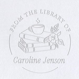 May include: A white embossed design featuring the text "FROM THE LIBRARY OF" in a circular pattern. Below the text is the name "Caroline Jenson". The design includes a stack of books, a steaming cup, and a floral element.