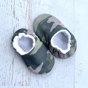 May include: A pair of baby booties in a camouflage pattern. The booties are primarily green, gray, and brown, with a soft white lining visible around the opening. The shoes are designed for comfort and warmth.