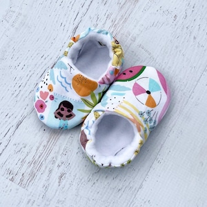 May include: A pair of baby shoes with a colorful summer-themed pattern. The shoes feature illustrations of a mermaid, watermelon, and beach elements on a white background. The interior is lined with white fabric.