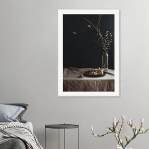 Könnte beinhalten: Gerahmte Stillleben-Fotografie mit dunkler, stimmungsvoller Ästhetik. Eine Glasflasche enthält getrocknete Zweige, und ein schwarzer Teller zeigt dekorative Objekte. Die Szene ist auf einem mit Leinen bedeckten Tisch vor dunklem Hintergrund aufgebaut.