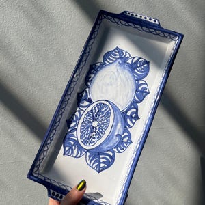 May include: A white ceramic rectangular serving tray with blue and white painted lemons and leaves. The tray has two blue handles on the sides.
