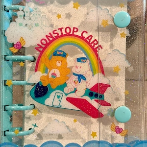 May include: A light blue glittery plastic binder featuring a graphic of two Care Bears in a plane with a rainbow and the text NONSTOP CARE. The cover has a scalloped cloud border, two circular snap closures, and a multi-ring spine mechanism.