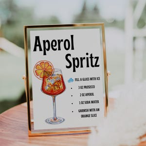 May include: Aperol Spritz recipe sign in a gold frame. The sign includes a watercolor illustration of the cocktail with an orange slice garnish. The recipe calls for prosecco, Aperol, and soda water.