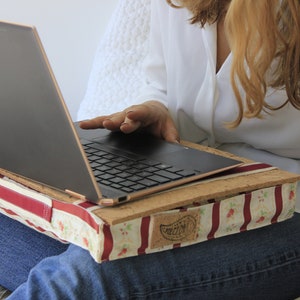 Può includere: Una donna sta usando un computer portatile su un cuscino per laptop a strisce rosse e bianche. Il cuscino ha un top in sughero e una maniglia in pelle marrone. Il laptop è nero con un logo HP argentato.