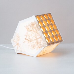 May include: White lamp with a geometric design and a wood panel with circular cutouts. The lamp is lit and casts a warm glow.