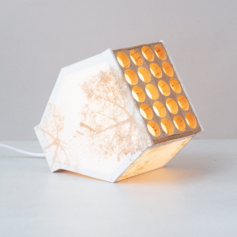 May include: White lamp with a geometric design and a wood panel with circular cutouts. The lamp is lit and casts a warm glow.