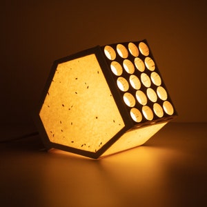 May include: A white paper lampshade with a wooden frame and a grid of circular cutouts. The lampshade is illuminated from within, casting a warm glow.