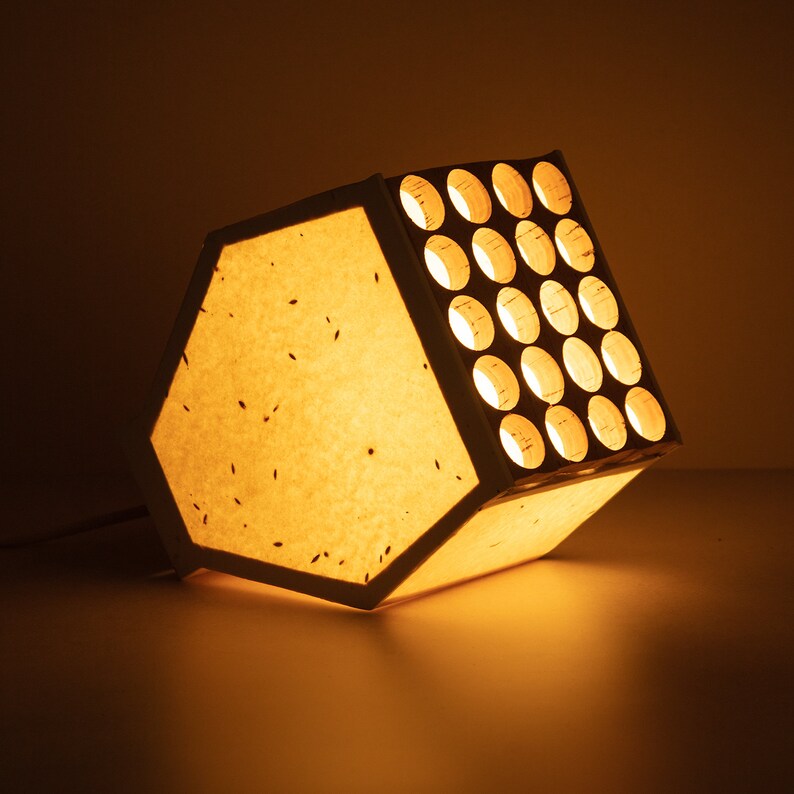 May include: A white paper lampshade with a wooden frame and a grid of circular cutouts. The lampshade is illuminated from within, casting a warm glow.