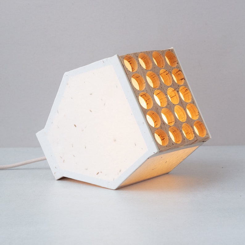 May include: A white table lamp with a honeycomb-like design on one side. The lamp is made of paper and has a warm, white light.