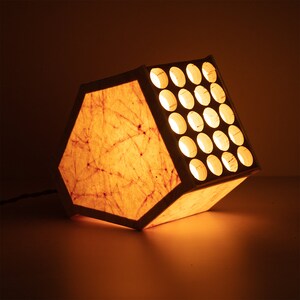 May include: A hexagonal paper lampshade with a light brown and white marbled pattern. The lampshade has a grid of round holes on one side, allowing light to shine through.