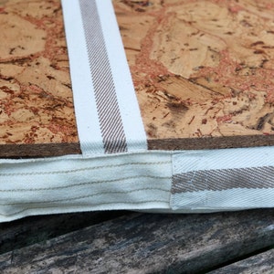 May include: A close-up of a cork board with a white and brown striped fabric border. The fabric is sewn around the edge of the cork board.