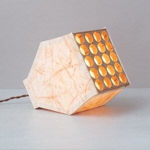May include: A white paper lampshade with a wooden frame featuring a grid of circular cutouts. The lampshade is lit from within, casting a warm glow.