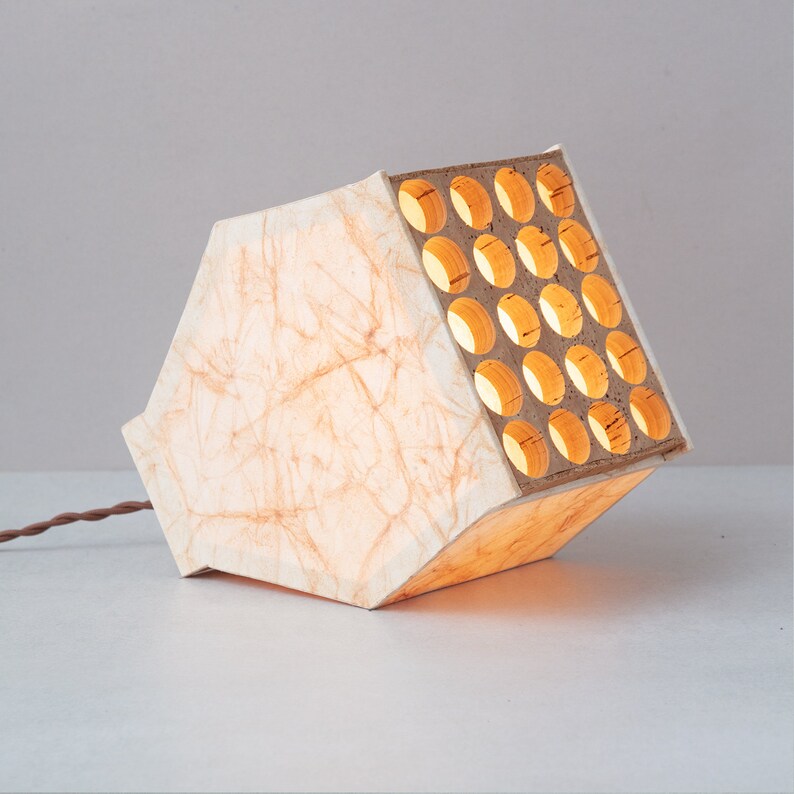 May include: A white paper lampshade with a wooden frame featuring a grid of circular cutouts. The lampshade is lit from within, casting a warm glow.