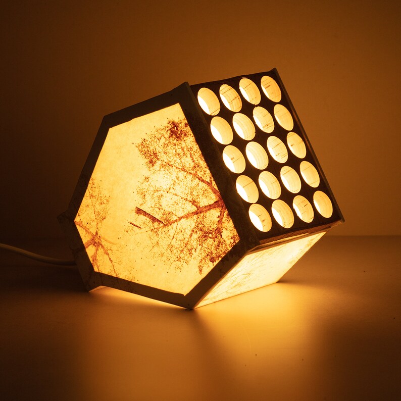 May include: A white lamp with a geometric design and a light brown pattern on the front. The lamp has a grid of round holes on the side.