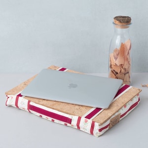 May include: A laptop computer sits on a cork and fabric laptop stand with a red and white striped pattern. The stand has a floral design and is sitting on a white surface. A glass jar filled with light brown snacks is in the background.