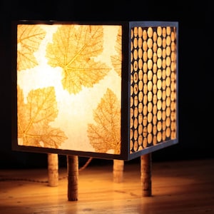 Op de afbeelding: Een vierkante lamp met een lichtbruin bladontwerp aan de voorkant en een kurkpatroon aan de zijkant. De lamp heeft drie houten poten.