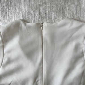 May include: A close-up of the back of a cream-colored garment. The fabric is smooth and appears to be a lightweight material. A silver zipper runs down the center of the back, providing a secure closure. The garment's neckline is visible.