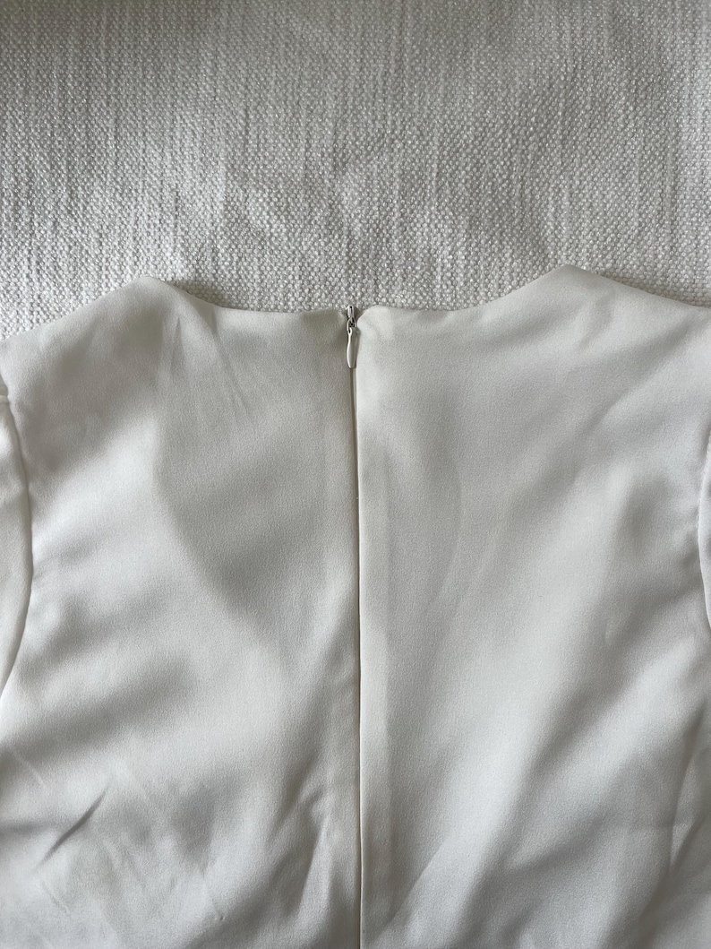 May include: A close-up of the back of a cream-colored garment. The fabric is smooth and appears to be a lightweight material. A silver zipper runs down the center of the back, providing a secure closure. The garment's neckline is visible.
