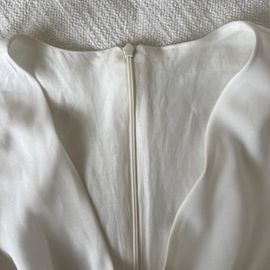 May include: Close-up of a white dress with a V-neckline and a zipper closure. The zipper is centered and runs vertically down the front. The fabric appears to be a soft, flowing material.
