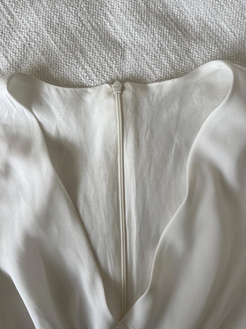 May include: Close-up of a white dress with a V-neckline and a zipper closure. The zipper is centered and runs vertically down the front. The fabric appears to be a soft, flowing material.