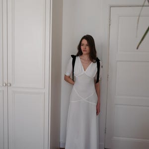 May include: A white, floor-length dress with a V-neckline and short sleeves. The dress features black shoulder ties and a fitted bodice with a flowing skirt. The dress is worn with black shoes.