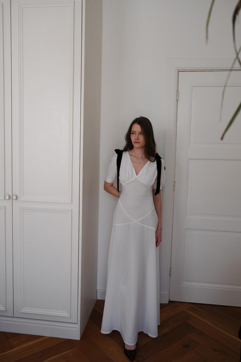 May include: A white, floor-length dress with a V-neckline and short sleeves. The dress features black shoulder ties and a fitted bodice with a flowing skirt. The dress is worn with black shoes.