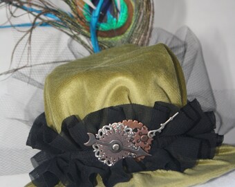May include: Olive green velvet hat with a black ruffled brim and a decorative metal gear and arrow accent. A peacock feather and blue accent feathers are attached to the top of the hat. The hat is a unique accessory.